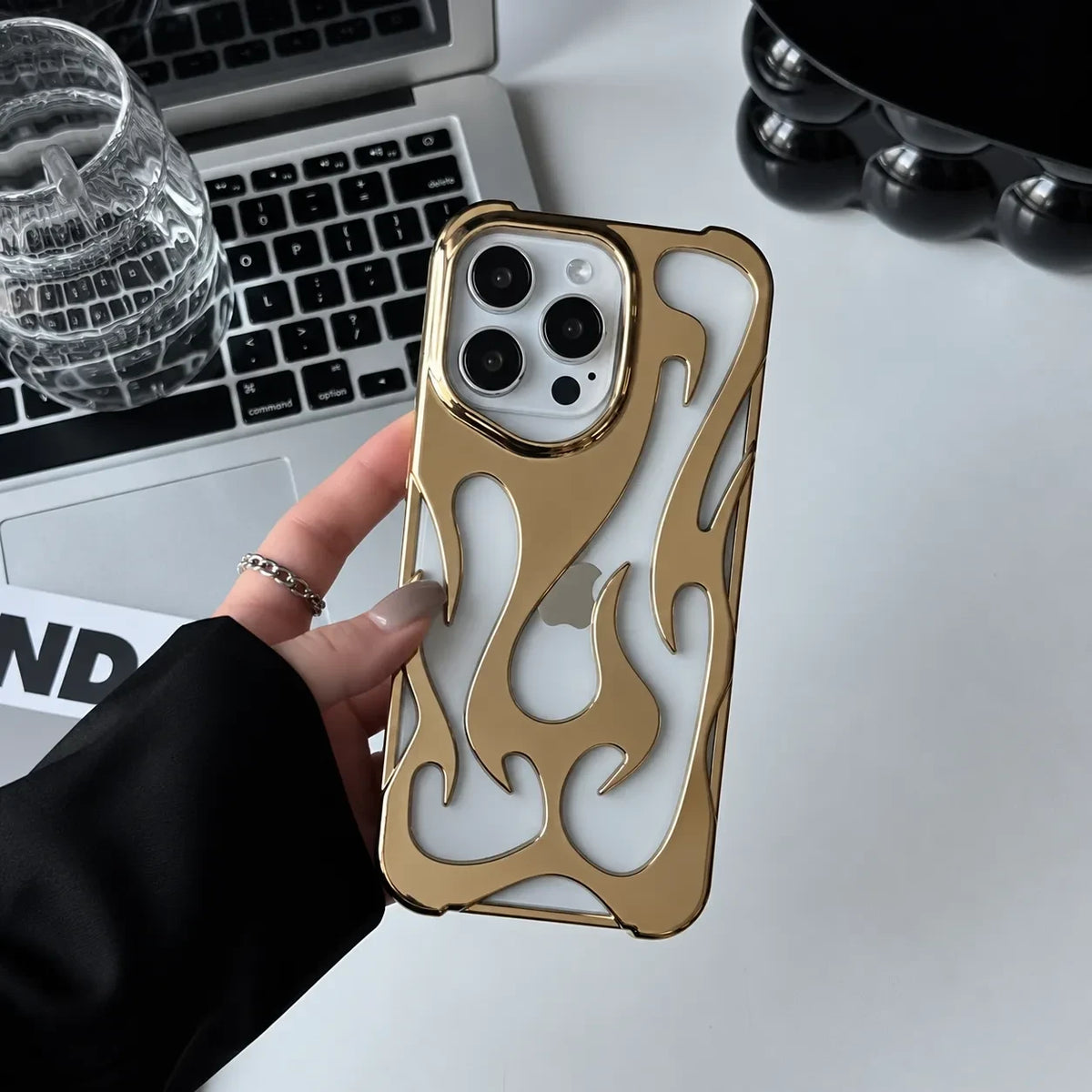 Zelpora gold chrome flame iPhone case with a translucent frosted back and raised metallic detailing on a white desk.